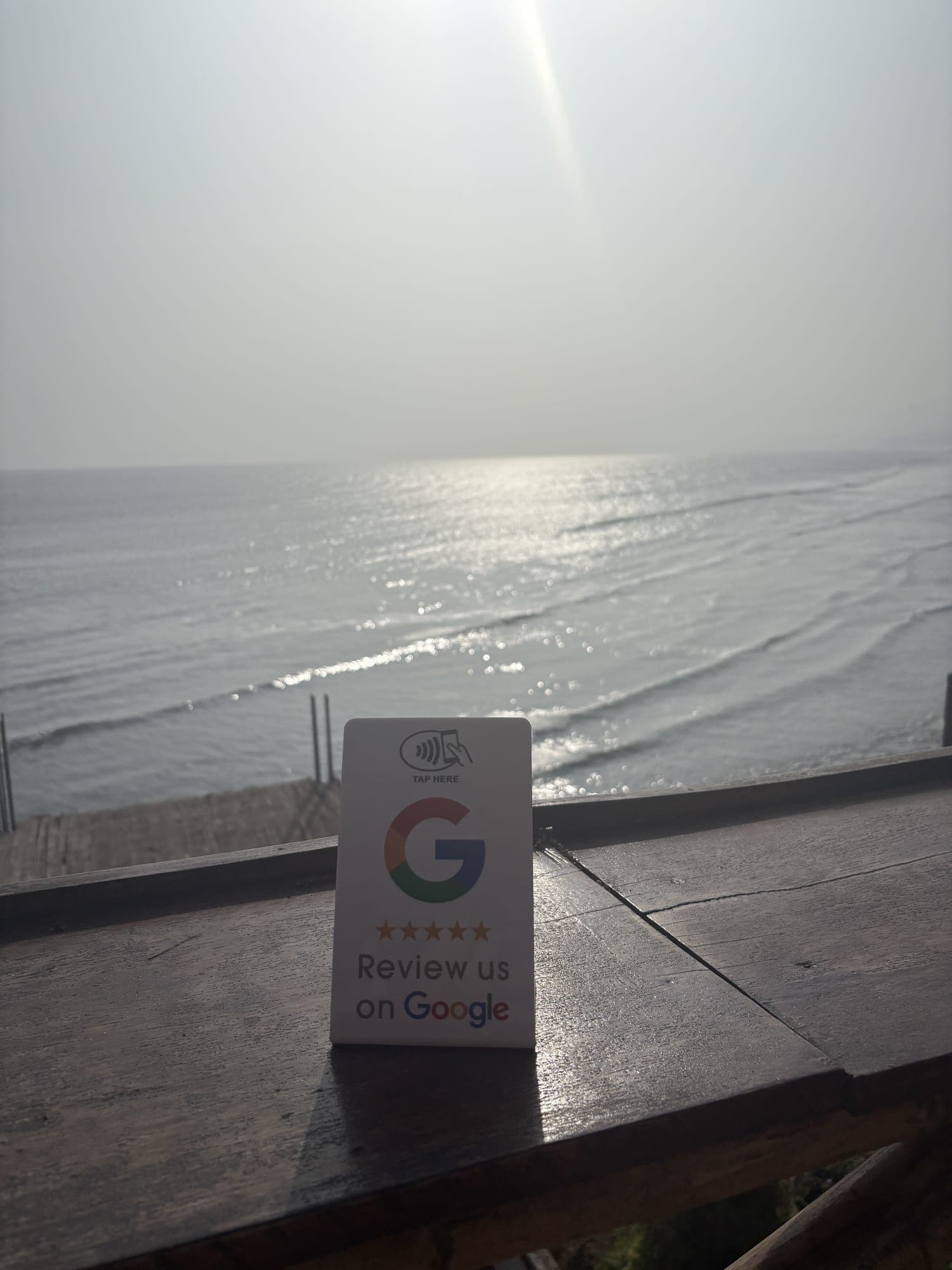 A Google review stand propped on a beachfront rail, with the sea and a low sun filling the frame behind it.