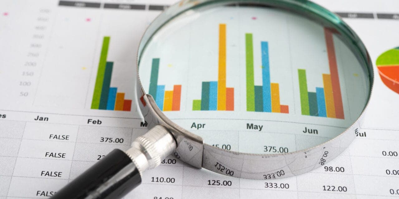 A magnifying glass held over printed bar charts, resting on a spreadsheet of monthly figures.