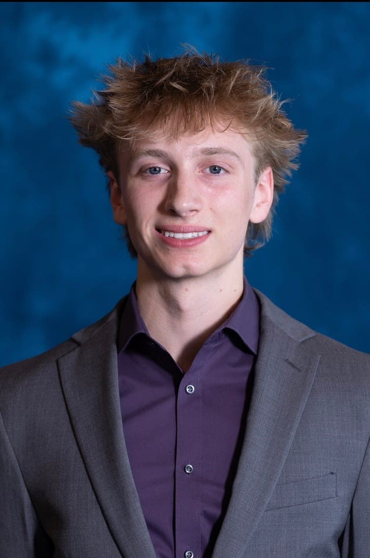 Head-and-shoulders studio portrait: light hair, a grey suit jacket over a purple shirt, against a mottled blue backdrop.