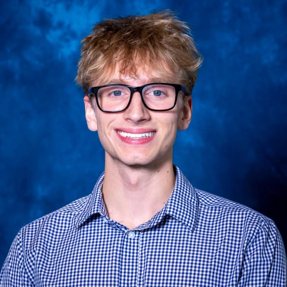 Head-and-shoulders studio portrait: short fair hair, black-framed glasses and a blue gingham shirt, against a mottled blue backdrop.