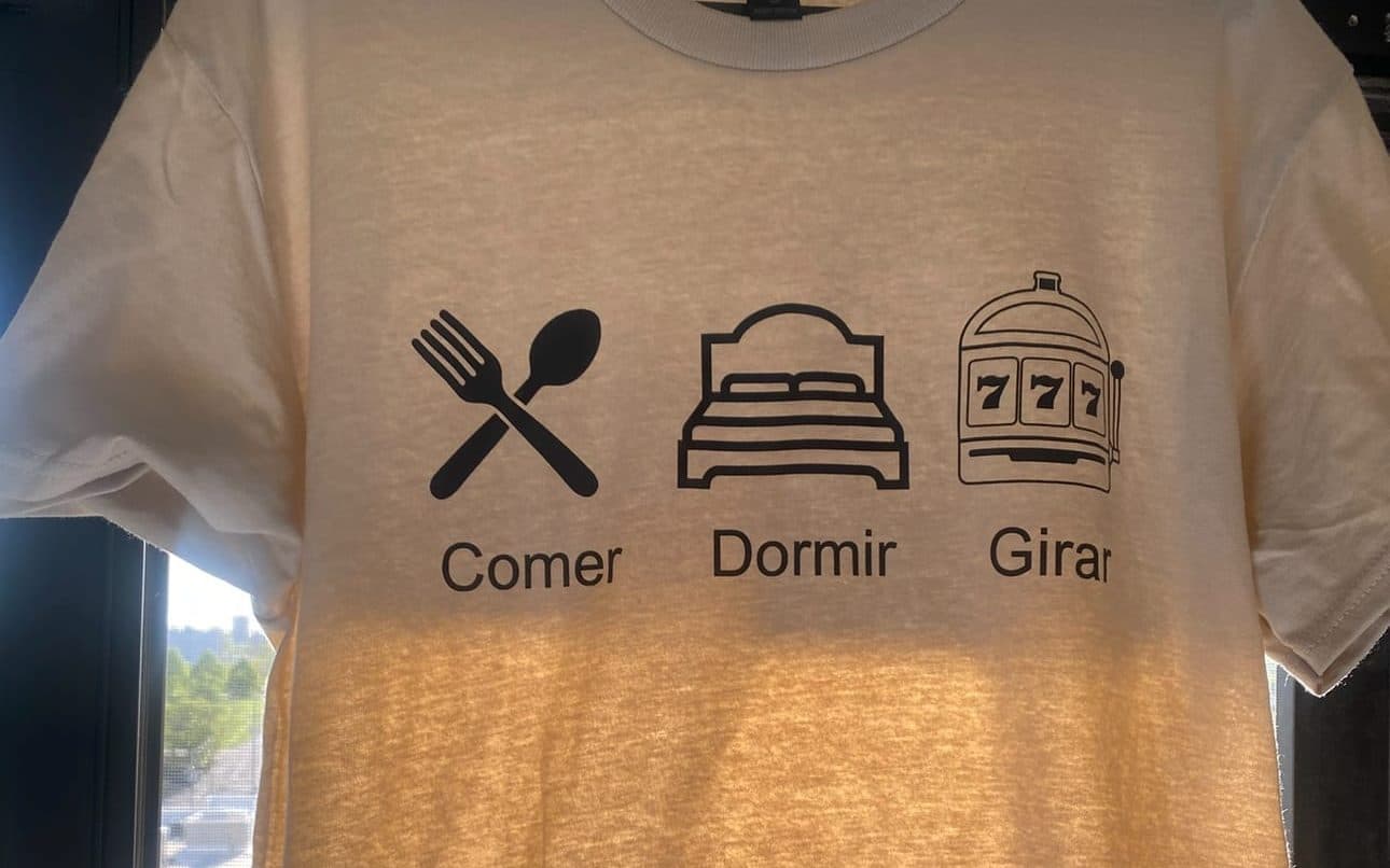 A sand-colored t-shirt printed with three icons and Spanish captions: a fork and spoon for "Comer", a bed for "Dormir", and a slot machine for "Girar".